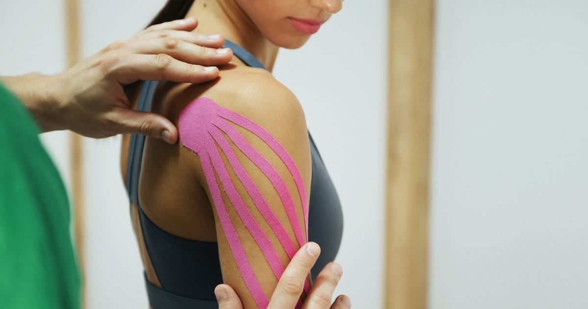 Kinesiologisches Taping als Hausbesuch in München – Physiotherapie Köpple Physiotherapeut bringt ein elastisches kinesiologisches Tape auf der Schulter einer Patientin an