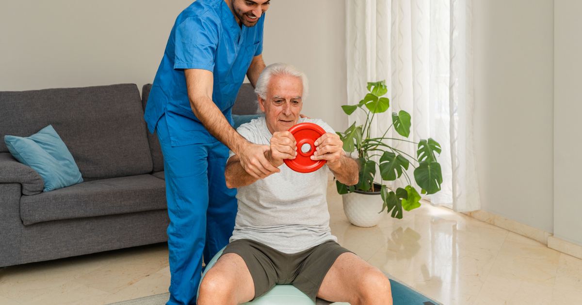 Senior trainiert unter Anleitung eines Physiotherapeuten mit einem Übungsgerät in seinem Wohnzimmer